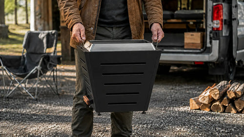Person carrying 20 inch slatted smokeless fire pit by built-in side handles at outdoor camping site with vehicle and firewood nearby, demonstrating the compact fire pit's portability for easy transport and placement in vehicles after proper cooling.