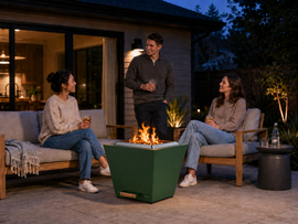 Three young adults relaxing around a green 25-inch smokeless fire pit on a modern backyard patio at dusk.