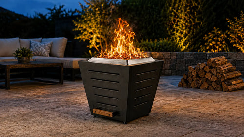 Legacy Heating 20-inch black slatted smokeless fire pit burning with low smoke on a stone patio beside stacked firewood at dusk.