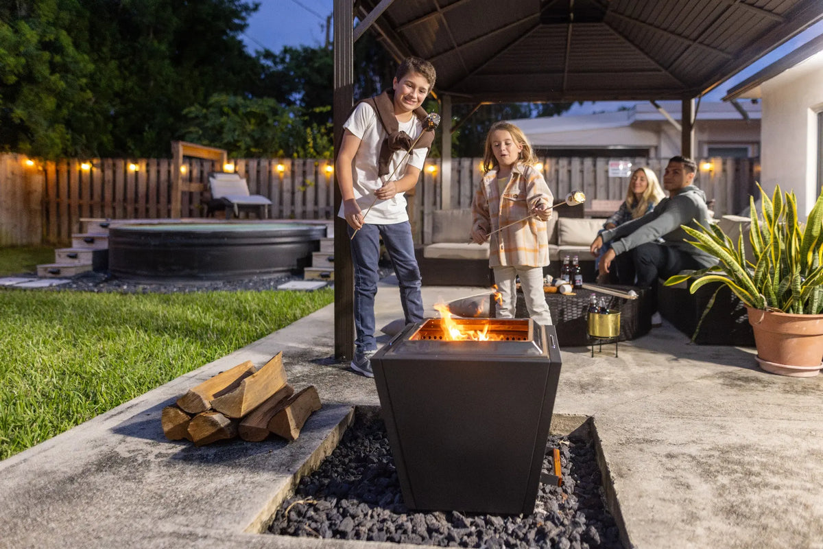 Children roast marshmallows over a black LEGACY HEATING 20-inch Flat Smokeless Fire Pit while adults relax on a covered backyard patio at dusk.