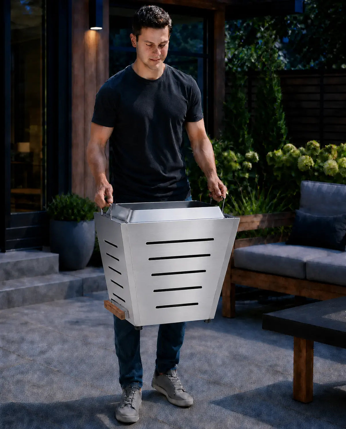 Young man easily carrying a LEGACY HEATING 25-inch stainless steel slatted smokeless fire pit using its two raised integrated stainless steel carry handles for convenient portability.