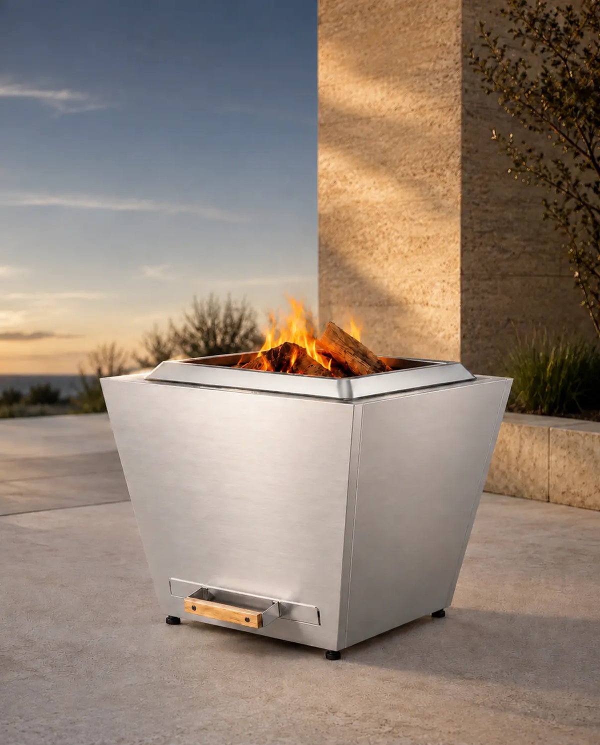 LEGACY HEATING 25 Inch Flat Smokeless Fire Pit in brushed stainless steel, featuring a tapered four-panel body, raised fire ring, and natural wood fire on a contemporary concrete and stone patio at sunset.